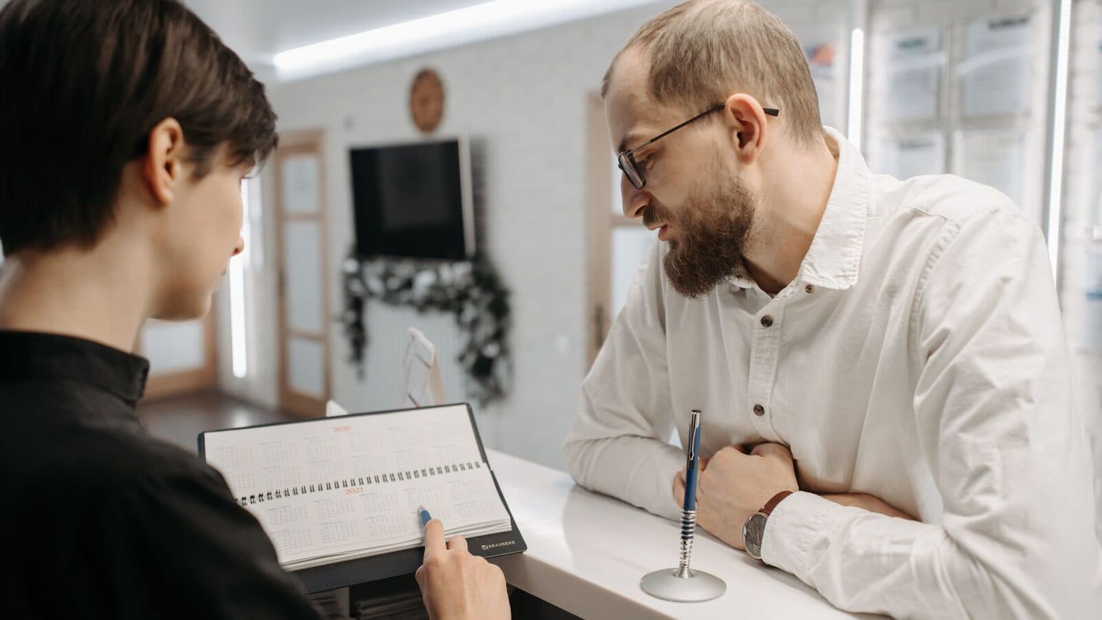 A receptionist and client converse over an appointment book at a clinic reception desk.